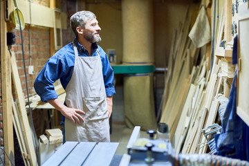 Portrait of bearded mature carpenter smiling looking at wood boards in workshop, copy space