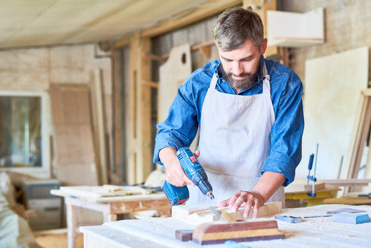 Portrait of mature bearded carpenter drilling wooden part while making furniture in modern workshop, copy space