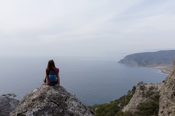 Fototapeta premium Rear view of a young woman sitting at the top of a mountain, looking into the distance on the top of mountain. Background is mountains and cloudy sky.