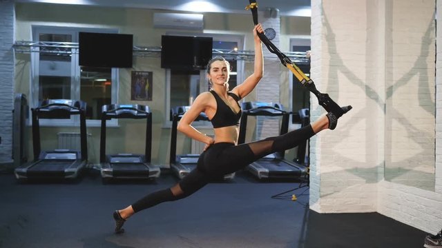 A young and pretty fitness trainer stretches the muscles of the legs with the help of a sports rubber band, twine, stretching legs with suspended arms, gym hd