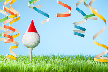 Concept for celebration events at a golf course or for a golf enthusiast with golf ball on tee wearing party hat and blow-out streamers coming down from top
