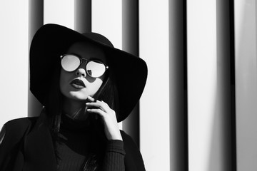 Fashion portrait of model in trendy hat and stylish glasses posing at the shutters. Black and white...