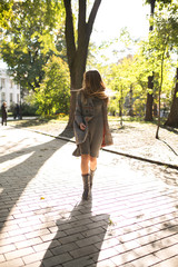 Elegant young woman with long hair in knit dress and warm coat walking in the park in sunny day