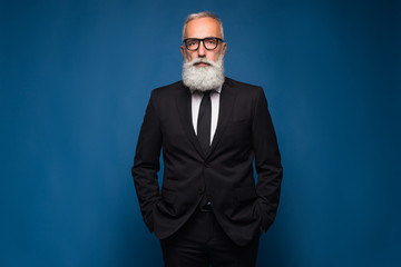 Confident senior bearded man in a suit and tie stand in front of blue isolated background. Fashion...
