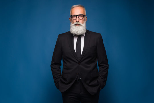 Confident senior bearded man in a suit and tie stand in front of blue isolated background. Fashion posing adult man in fashion glasses and suit