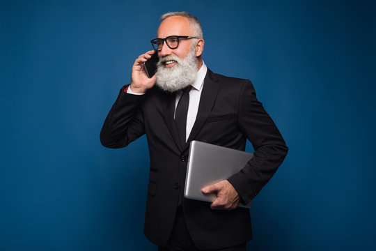 Busy bearded senior and business man staying with notebook and mobile phone isolated on a blue background. Working business man talk on the phone in straight suit and glasses