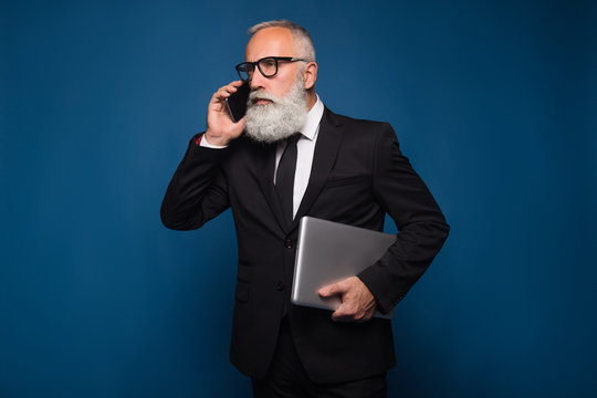 Busy bearded serious senior and business man staying with notebook and mobile phone isolated on a blue background. Working business man talk on the phone in straight suit and glasses.