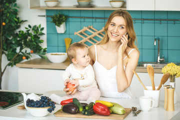 Young mother smiling, cooking and playing with her baby daughter in a modern kitchen. Using phone. Healthy food concept. Working at home.