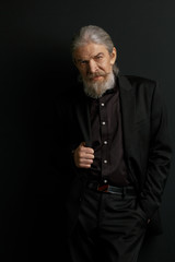 Stylish aged man with lonf hair and beard standing in studio against black wall. Portrait of very old wrinkled male.