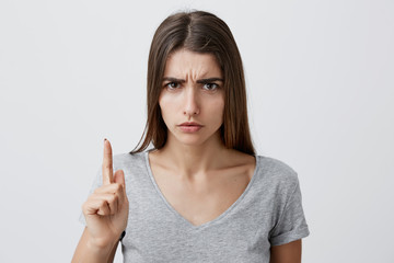 Portrait of beautiful young caucasian student girl with long brown hair in casual gray t-shirt, gesticulating with index finger, looking with angry expression at little kid in zoo, showing not to
