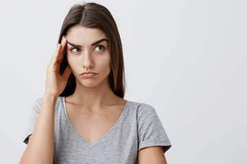 Close up of young handsome serious caucasian girl with long brown hair in stylish gray t-shirt touching forehead with hand, looking aside with tired face expression after long day in university.