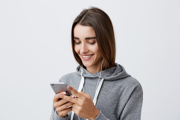 Close up of young attractive happy caucasian female student with long dark hair in gray hoodie laughing, looking funny video from party on phone, listening to favorite song with earphones.