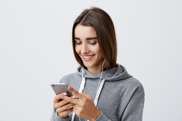 Naklejka premium Close up of young attractive happy caucasian female student with long dark hair in gray hoodie laughing, looking funny video from party on phone, listening to favorite song with earphones.