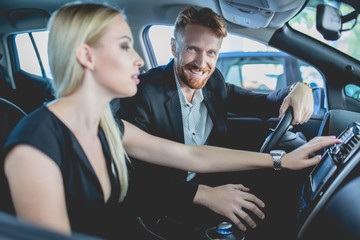 Pretty couple in auto showroom choosing car. Man and woman inside electric car, examining...