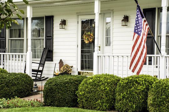 white colonial home decorated for fall with American flag flying from the front porch with rocking chairs