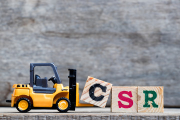 Yellow plastic forklift hold letter C to complete word CSR (Abbreviation from corporate social responsibility) on wood background