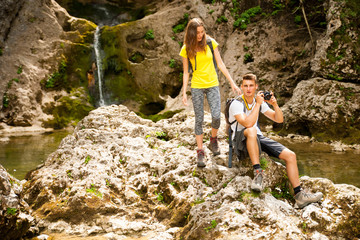 Naklejka na ściany i meble active young couole hiking in nature resting near waterfalll and taking pictures with camera