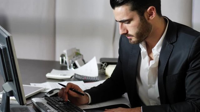 Tired bored young businessman sitting at his desk in front of his computer with his chin resting on his hand, sleepy