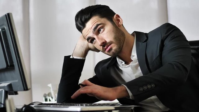 Tired bored young businessman sitting at his desk in front of his computer with his chin resting on his hand, sleepy