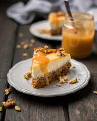 Carrot nut vanilla cheesecake with salty homemade caramel on a white plate on a dark wooden vintage background. Two pieces of homemade cheesecake. Caramel sauce in a jar. - obrazy, fototapety, plakaty