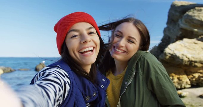 Two attractive young women making smiled selfies with victory signs on the rocky sea background. Cold sunny weather. Outside. POV. Close up
