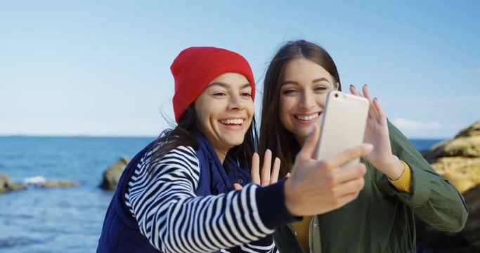 Two attractive female friends having video chatting on the smart phone on the rocky sea background. Cold sunny weather. Outside.