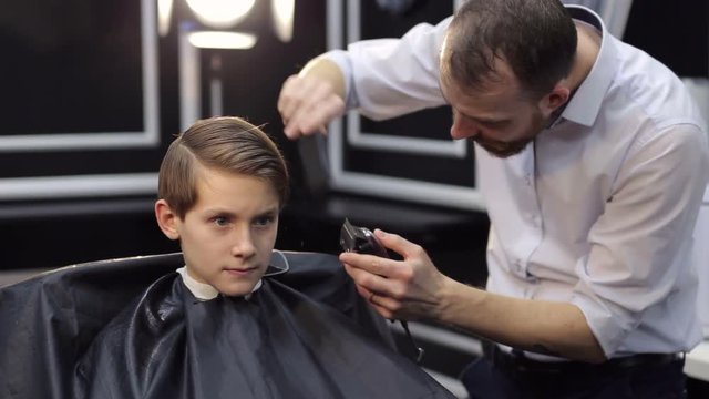 Professional hairdresser working in a beauty salon with a boy teenager. Male hairdresser cuts a young boy in a barbershop. Middle shot.
