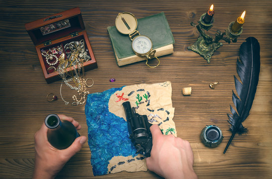 Treasure map, compass and feather pen on wooden table. Treasure hunt. Adventurer man holding in one hand handgun and showing with it on place with gold on treasure map and rum bottle in another. - Powered by Adobe
