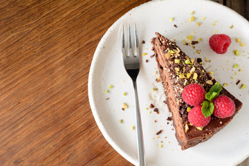 Slice of chocolate cake topped with raspberries, pistachio nut and mint leaf on white plate. Copy space for text. Top view