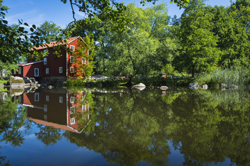 Beautiful picture of a Swedish watermill where the quaint café Ekenas Kvarn is situated. A colorful photo of the typical red mill with green trees and a mirror-lik pond reflecting the gorgeous surroun
