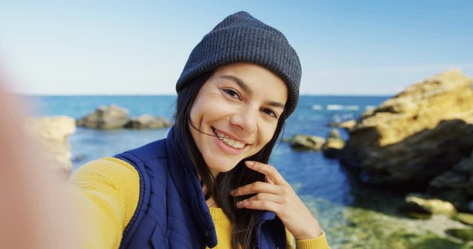 Attractive brunette woman making nice selfies on the rocky sea beach. Cold sunny weather. Outdoors. POV. Close up. Portrait