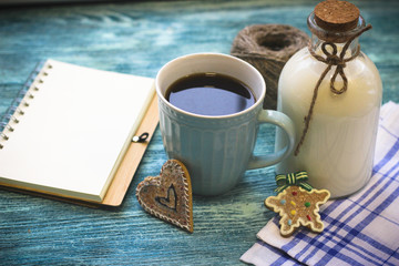 Festive still life with a bottle of milk, a cup of freshly brewed fragrant coffee, biscuits, a skein of yarn, a notepad, a checkered tablecloth on a turquoise wooden table, a light background