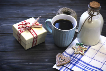 Festive still life with a bottle of milk, a cup of freshly brewed fragrant coffee, biscuits, a skein of yarn, a gift box, a checkered tablecloth on a gray wooden table, a dark background