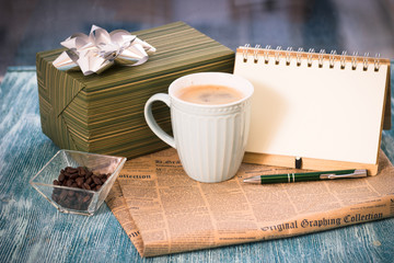 Festive still life with a cappuccino cup, gift box, a vase with coffee beans, a notebook with a pen, a newspaper on a turquoise wooden table, a country background on a sunny day