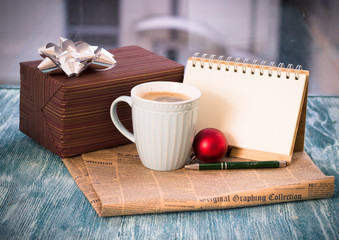 Festive still life with a cappuccino cup, gift brown box, red Christmas-tree ball, notebook with pen, newspaper on a turquoise wooden table, country background on a sunny day