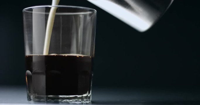 Adding foamed milk into a shot of espresso in a clear shot glass isolated on dark background