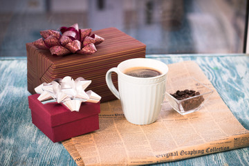 Festive still life with a cappuccino cup, two gift boxes with bows, a vase with coffee beans, a newspaper on a turquoise wooden table, a country background on a sunny day
