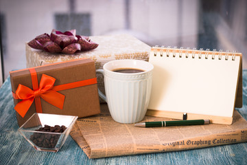 Festive still life with a cup of freshly brewed coffee, two gifts in brown and golden boxes, a vase with coffee beans, a notebook with a pen, a newspaper on a turquoise wooden table, a country