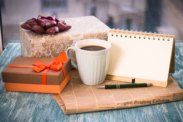 Festive still life with a cup of freshly brewed coffee, two gifts in brown and golden boxes, a notebook with a pen, a newspaper on a turquoise wooden table, a country background on a sunny day