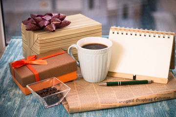 Festive still life with a cup of freshly brewed coffee, two gifts in brown and light boxes, a vase with coffee beans, a notebook with a pen, a newspaper on a turquoise wooden table, a country