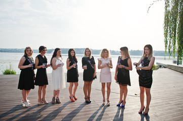 Obraz premium Group of 8 girls wear on black and 2 brides at hen party against sunny beach drinking champagne.