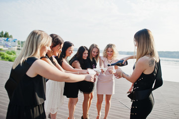 Group of 8 girls wear on black and 2 brides at hen party against sunny beach drinking champagne.