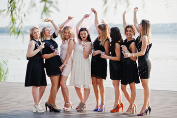 Group of 8 girls wear on black and 2 brides at hen party against sunny beach drinking champagne.