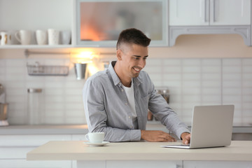 Naklejka premium Young man using laptop at table in kitchen