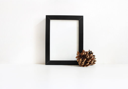 Black vertical blank wooden frame mockup with pine cones lying on the white table. Poster product design. Styled stock feminine photography. Home decor. Christmas winter concept.