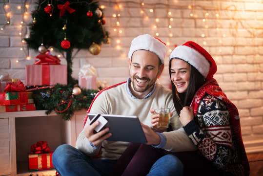 Lovely charming couple sitting on a floor with Santa hats hugged while shopping online on a tablet and drinking a tea for Chrismas holidays.