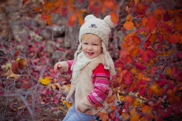 Cute young russian baby girl stylish dressed in warm white fur handmade jacket blue jeans boots and hooked hat teddy bear posing in autumn colorful forest pathway Face with pink cheek