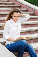 Young woman sitting on stairs and reading a book. Beautiful european girl studying a subject with book