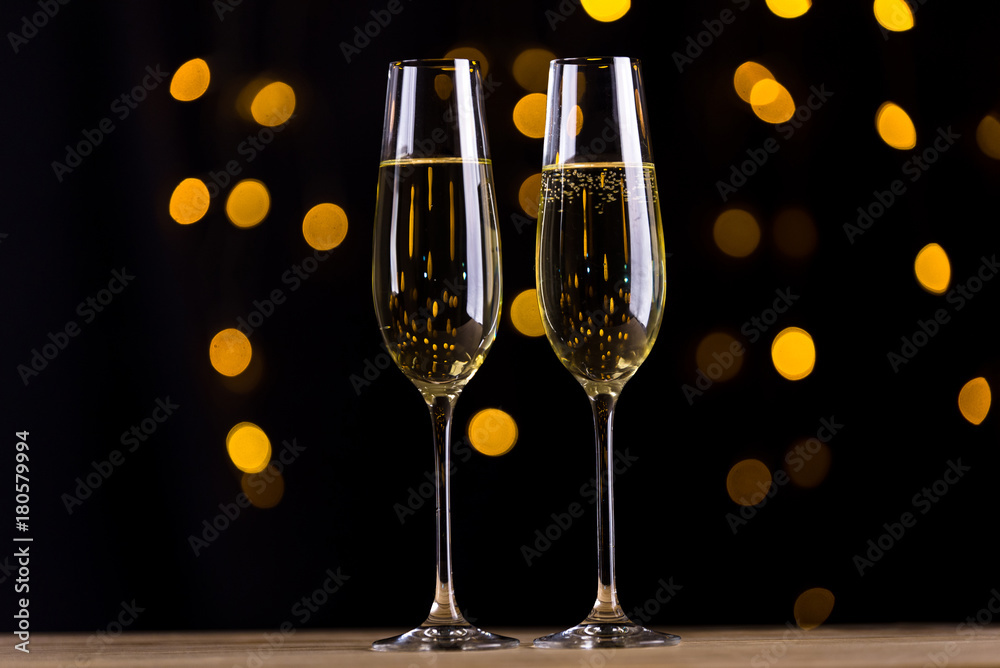 Wall mural Two champagne glasses on black background and bokeh garland light beams and glitters