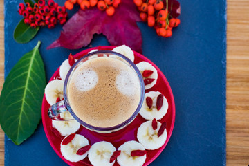 Transparent cup of coffee with foam on a red saucer with pieces of banana and goji berries.